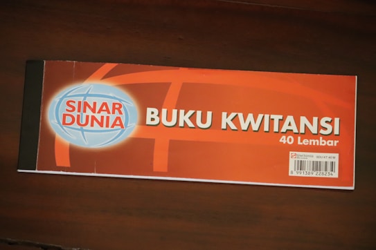A receipt book labeled 'Buku Kwitansi' with 40 sheets, featuring the brand 'Sinar Dunia'. The cover is predominantly orange with a blue and white globe logo on the left. A barcode is visible on the bottom right corner, along with some additional information.