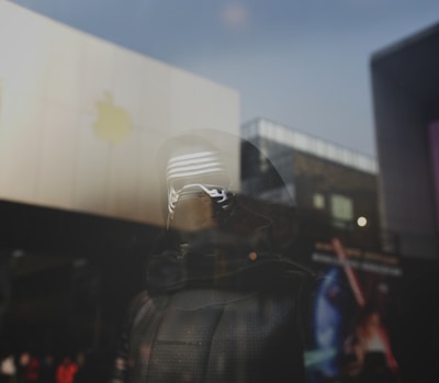 A glass reflection shows a character wearing a dark hood and mask with futuristic elements. The background includes modern architecture and the faint logo of a large tech company visible on a building. The scene appears to blend real-world and cinematic elements.
