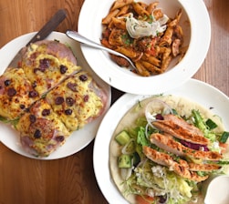 A table with three white plates of different dishes. The first plate contains slices of pizza-like bread topped with melted cheese, olives, and herbs. The second plate has a serving of penne pasta mixed with a tomato-based sauce and garnished with herbs and onions. The third plate features a salad with grilled chicken slices, mixed greens, cucumbers, onions, and a side of dressing, all served on flatbread.
