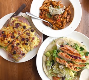 A table with three white plates of different dishes. The first plate contains slices of pizza-like bread topped with melted cheese, olives, and herbs. The second plate has a serving of penne pasta mixed with a tomato-based sauce and garnished with herbs and onions. The third plate features a salad with grilled chicken slices, mixed greens, cucumbers, onions, and a side of dressing, all served on flatbread.