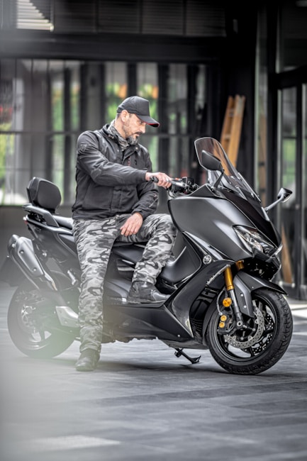 A man in a black jacket and camouflage pants is sitting on a black scooter in an indoor setting with a faint glow around the edges. He is wearing a black cap and appears to be adjusting something on the scooter's handlebar. The background features a modern industrial-style room with large windows.