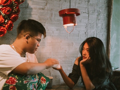 A woman and a man sit facing each other in a cozy, dimly lit environment. The woman rests her chin on her hand and looks thoughtfully towards the man, who is holding a napkin or tissue. A wall decorated with red roses and an industrial-style lamp provide a warm ambiance.