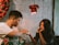 A woman and a man sit facing each other in a cozy, dimly lit environment. The woman rests her chin on her hand and looks thoughtfully towards the man, who is holding a napkin or tissue. A wall decorated with red roses and an industrial-style lamp provide a warm ambiance.