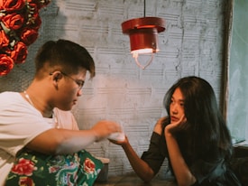 A woman and a man sit facing each other in a cozy, dimly lit environment. The woman rests her chin on her hand and looks thoughtfully towards the man, who is holding a napkin or tissue. A wall decorated with red roses and an industrial-style lamp provide a warm ambiance.