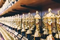 A collection of gold-colored statues resembling awards lined up on shelves. Each statue features a standing figure holding an object, set against a perspective view that emphasizes the rows. The lighting highlights the shiny, reflective surfaces of the trophies.