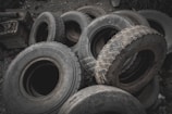A pile of worn-out tires with various tread patterns stacked in an outdoor setting, surrounded by rocks and debris. The tires appear used and are arranged haphazardly.