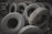 A pile of worn-out tires with various tread patterns stacked in an outdoor setting, surrounded by rocks and debris. The tires appear used and are arranged haphazardly.