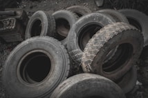 A pile of worn-out tires with various tread patterns stacked in an outdoor setting, surrounded by rocks and debris. The tires appear used and are arranged haphazardly.