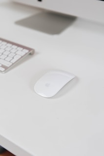 A sleek and modern white computer mouse is placed on a white desk alongside a wireless keyboard. Part of a monitor stand is visible in the background, creating a clean and minimalistic workspace setting.