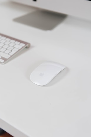 A sleek and modern white computer mouse is placed on a white desk alongside a wireless keyboard. Part of a monitor stand is visible in the background, creating a clean and minimalistic workspace setting.