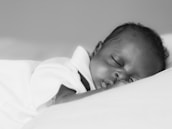 A peaceful close-up of a sleeping baby with eyes gently closed against a soft pillow. The baby appears serene and comfortable in this black and white photograph.
