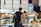 A man with a backpack stands at a cheese counter in a market. Various types of cheeses are displayed, and a vendor is assisting him. Signs in German describe the cheese selections with prices.