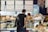 A man with a backpack stands at a cheese counter in a market. Various types of cheeses are displayed, and a vendor is assisting him. Signs in German describe the cheese selections with prices.