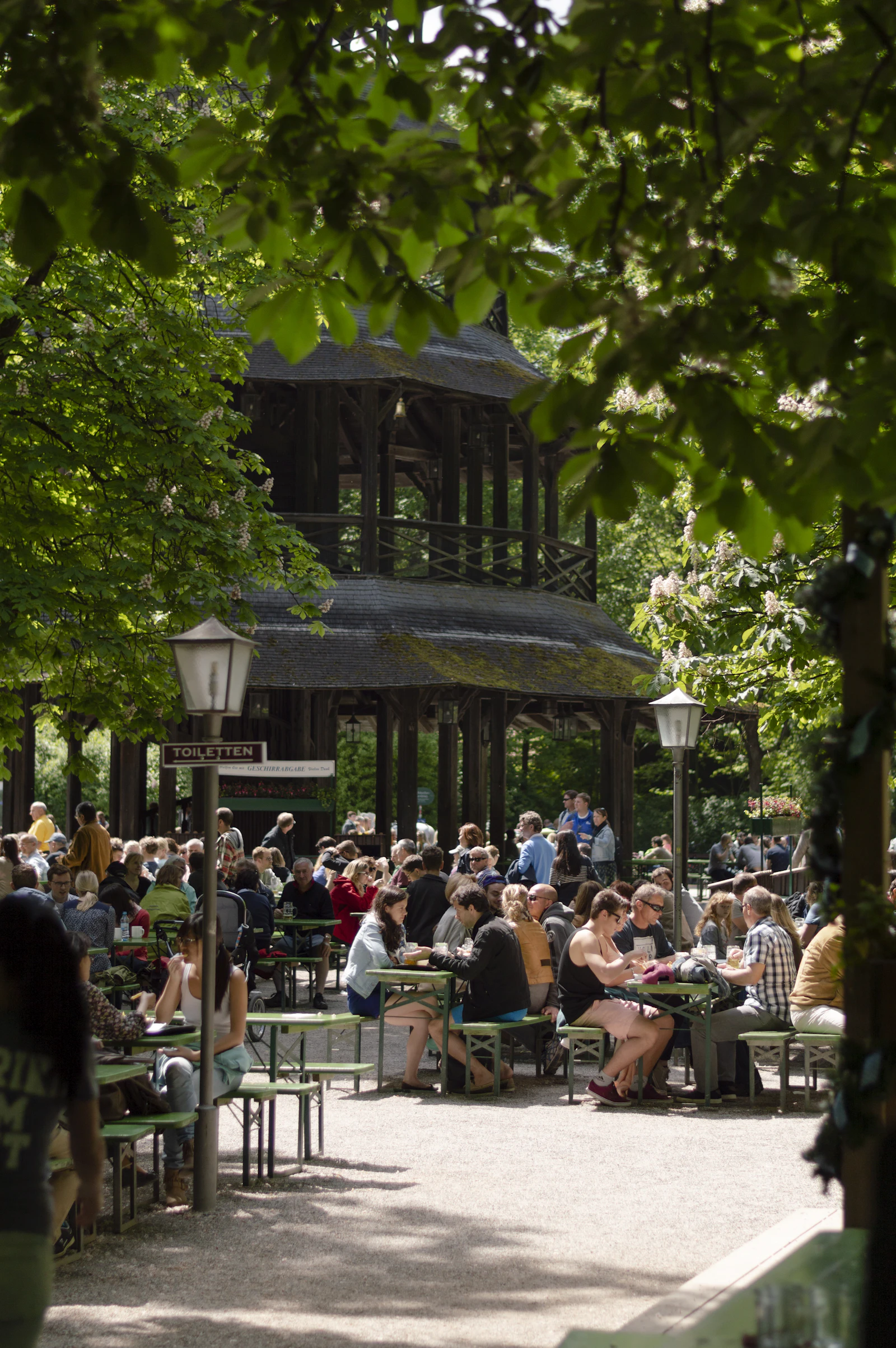 People sit at long green benches beneath a leafy canopy in an outdoor beer garden.
