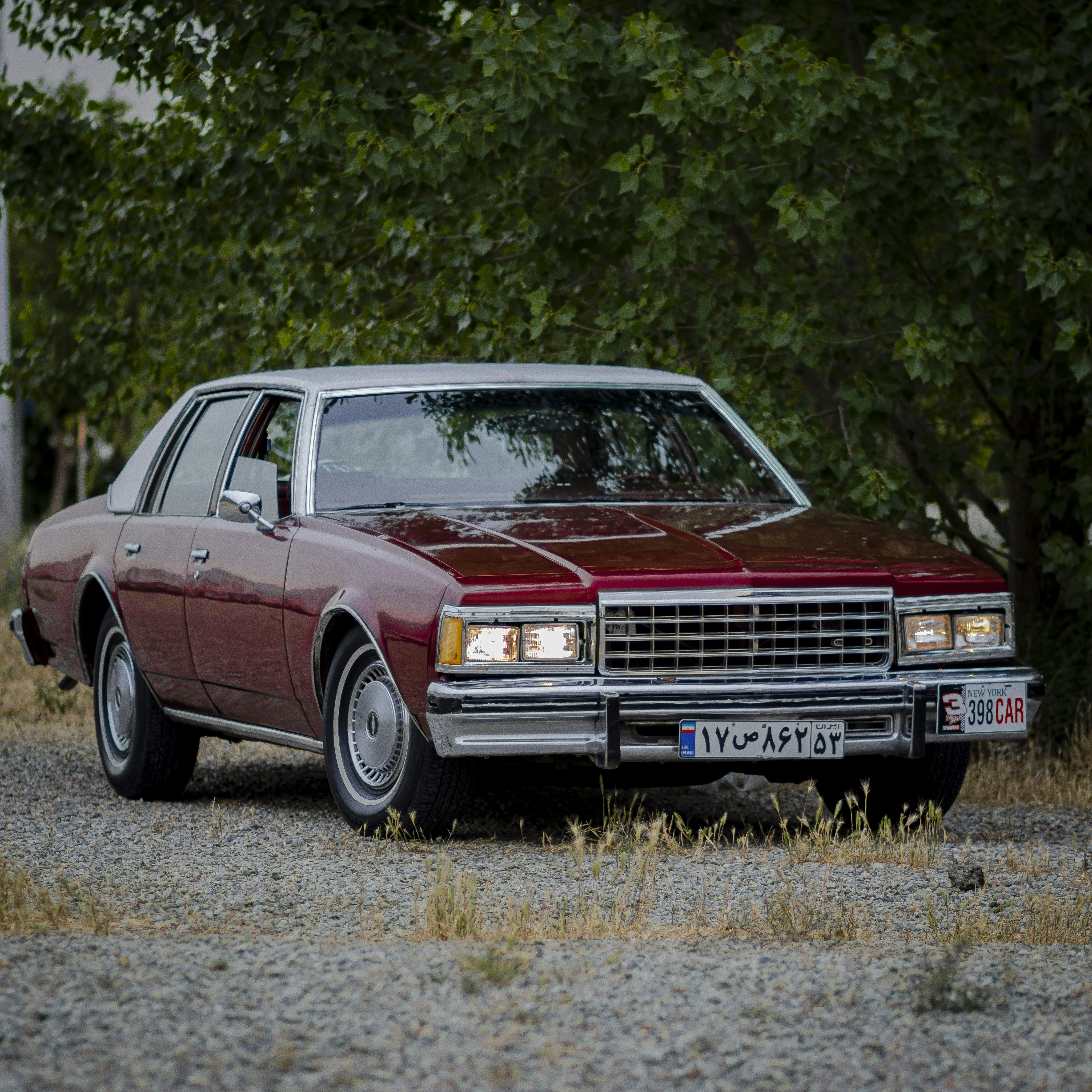 A vintage red sedan rests on a gravel surface beneath a canopy of green trees, its chrome grille catching the light in this outdoor photograph.
