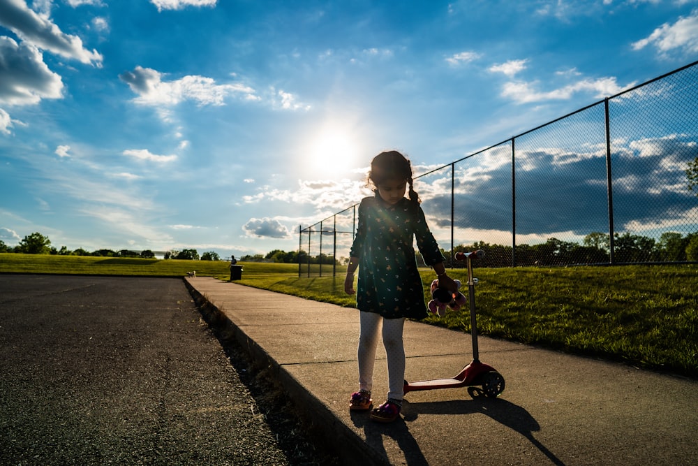 girl in green dress holding kick scooter on gray concrete pathway during daytime