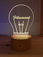 A wooden base supports an acrylic light resembling a lightbulb with the word 'Filament' inscribed on it. The design is softly illuminated, creating a warm, cozy glow.