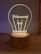 A wooden base supports an acrylic light resembling a lightbulb with the word 'Filament' inscribed on it. The design is softly illuminated, creating a warm, cozy glow.