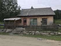 A rustic house with a thatched roof and wooden front is situated in a rural setting. The building has wooden shutters and a faded red door. Surrounding the house is a low wooden fence made of thin branches, and a cartwheel is visible on the left side. The area is framed by a gravel path and a line of stones, with green trees and a grassy field in the background.