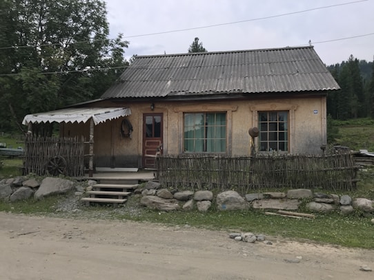 A rustic house with a thatched roof and wooden front is situated in a rural setting. The building has wooden shutters and a faded red door. Surrounding the house is a low wooden fence made of thin branches, and a cartwheel is visible on the left side. The area is framed by a gravel path and a line of stones, with green trees and a grassy field in the background.
