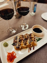 There is a rectangular white plate with a gourmet dish featuring sauce-drizzled food pieces, possibly sushi or appetizers, garnished with green and red condiments. A small bowl of dipping sauce is on the side. In the background are two half-filled glasses of red wine on a grey tablecloth.