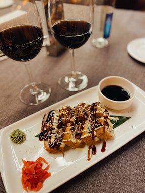 There is a rectangular white plate with a gourmet dish featuring sauce-drizzled food pieces, possibly sushi or appetizers, garnished with green and red condiments. A small bowl of dipping sauce is on the side. In the background are two half-filled glasses of red wine on a grey tablecloth.