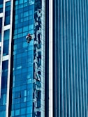 A person wearing a red helmet is suspended on ropes, cleaning or inspecting the glass windows of a modern high-rise building. The building features a grid of reflective blue glass panels, and the sunlight creates intricate patterns and shadows on the surface.