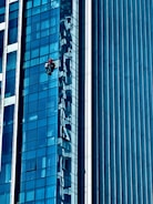 A person wearing a red helmet is suspended on ropes, cleaning or inspecting the glass windows of a modern high-rise building. The building features a grid of reflective blue glass panels, and the sunlight creates intricate patterns and shadows on the surface.