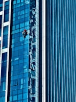A person wearing a red helmet is suspended on ropes, cleaning or inspecting the glass windows of a modern high-rise building. The building features a grid of reflective blue glass panels, and the sunlight creates intricate patterns and shadows on the surface.