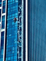 A person wearing a red helmet is suspended on ropes, cleaning or inspecting the glass windows of a modern high-rise building. The building features a grid of reflective blue glass panels, and the sunlight creates intricate patterns and shadows on the surface.
