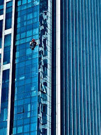 A person wearing a red helmet is suspended on ropes, cleaning or inspecting the glass windows of a modern high-rise building. The building features a grid of reflective blue glass panels, and the sunlight creates intricate patterns and shadows on the surface.