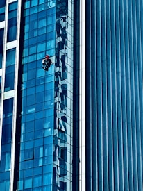 A person wearing a red helmet is suspended on ropes, cleaning or inspecting the glass windows of a modern high-rise building. The building features a grid of reflective blue glass panels, and the sunlight creates intricate patterns and shadows on the surface.