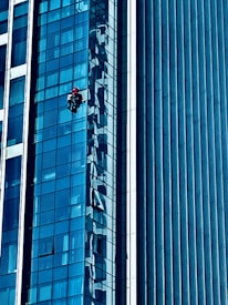 A person wearing a red helmet is suspended on ropes, cleaning or inspecting the glass windows of a modern high-rise building. The building features a grid of reflective blue glass panels, and the sunlight creates intricate patterns and shadows on the surface.