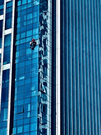 A person wearing a red helmet is suspended on ropes, cleaning or inspecting the glass windows of a modern high-rise building. The building features a grid of reflective blue glass panels, and the sunlight creates intricate patterns and shadows on the surface.