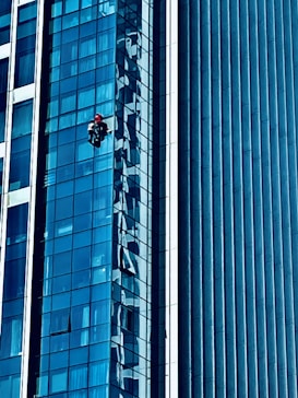 A person wearing a red helmet is suspended on ropes, cleaning or inspecting the glass windows of a modern high-rise building. The building features a grid of reflective blue glass panels, and the sunlight creates intricate patterns and shadows on the surface.