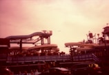 A waterslide park with multiple slides and structures. People can be seen on the platforms and slides, while a seagull flies overhead. Several cars are parked in the foreground. The lighting gives the image a warm, reddish tint.
