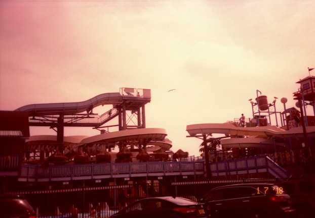 A waterslide park with multiple slides and structures. People can be seen on the platforms and slides, while a seagull flies overhead. Several cars are parked in the foreground. The lighting gives the image a warm, reddish tint.