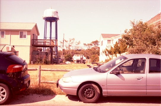 A residential neighborhood featuring several cars parked along a street. In the background, a tall water tower stands surrounded by houses and trees. The scene is set on a sunny day, casting shadows from the structures and vegetation.