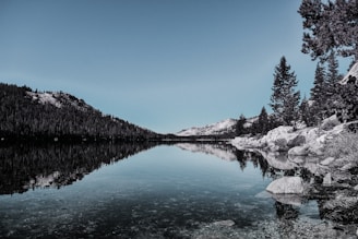 A serene mountain lake reflecting the surrounding forest, with a single figure standing quietly at the shore.