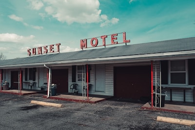 A vintage motel with a faded red sign spelling 'SUNSET MOTEL' sits under a blue sky with scattered clouds. The building features a gray roof with red and black accents, including poles and some furniture. Several windows and doors are visible along the front, with small patios outside the rooms. A rough asphalt parking lot extends in front of the building.