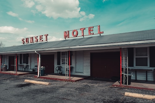 A vintage motel with a faded red sign spelling 'SUNSET MOTEL' sits under a blue sky with scattered clouds. The building features a gray roof with red and black accents, including poles and some furniture. Several windows and doors are visible along the front, with small patios outside the rooms. A rough asphalt parking lot extends in front of the building.
