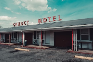A vintage motel with a faded red sign spelling 'SUNSET MOTEL' sits under a blue sky with scattered clouds. The building features a gray roof with red and black accents, including poles and some furniture. Several windows and doors are visible along the front, with small patios outside the rooms. A rough asphalt parking lot extends in front of the building.