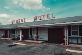 A vintage motel with a faded red sign spelling 'SUNSET MOTEL' sits under a blue sky with scattered clouds. The building features a gray roof with red and black accents, including poles and some furniture. Several windows and doors are visible along the front, with small patios outside the rooms. A rough asphalt parking lot extends in front of the building.