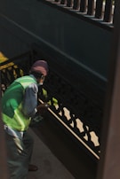 A person wearing a green vest and a brown hat appears to be cleaning or painting a darkly-colored railing on a staircase. The light creates shadows on the walls, and the person is holding a small object in their hand.