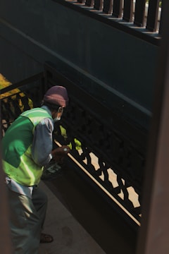A person wearing a green vest and a brown hat appears to be cleaning or painting a darkly-colored railing on a staircase. The light creates shadows on the walls, and the person is holding a small object in their hand.
