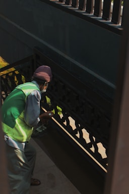 A person wearing a green vest and a brown hat appears to be cleaning or painting a darkly-colored railing on a staircase. The light creates shadows on the walls, and the person is holding a small object in their hand.
