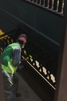 A person wearing a green vest and a brown hat appears to be cleaning or painting a darkly-colored railing on a staircase. The light creates shadows on the walls, and the person is holding a small object in their hand.