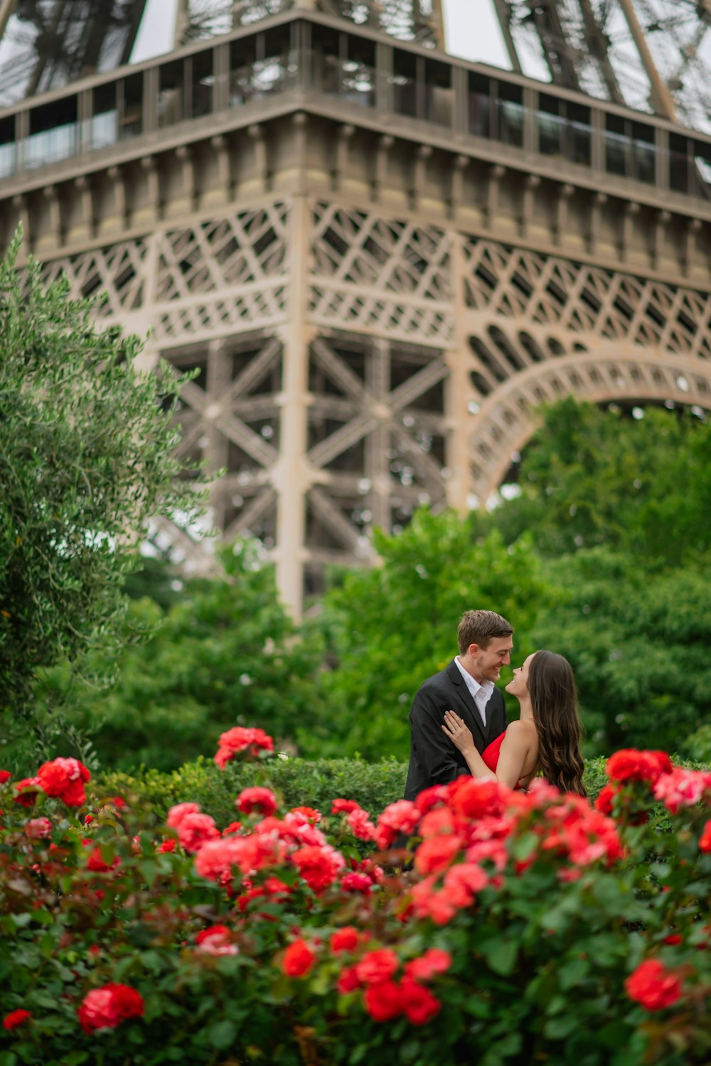 man in black suit standing beside woman in black dress surrounded by red flowers