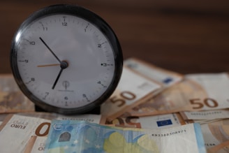 An analog clock with a black frame and white face is placed on top of scattered euro banknotes, including 50 and 10 euro bills, on a dark wooden surface.