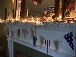A mantelpiece adorned with string lights and decorative garlands. American flags and star-themed decorations are prominently displayed, suggesting a patriotic theme. Candles and framed art pieces are also part of the arrangement, adding a cozy and festive atmosphere.